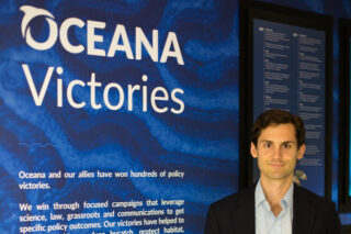 Russell Grandin (MBA '20) stands in front of a wall detailing the impacts of Oceana’s many policy victories