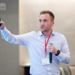 a man standing and speaking into a microphone as he gestures and looks at a screen to his right.