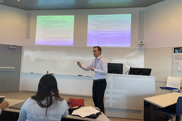 a man staking, speaking, and gesturing at the front of a classroom with slides on screens in the background.