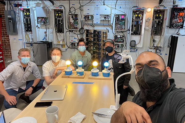 4 men and 1 woman wearing face masks are seated at a table with boards of 4 brightly lit light bulbs in front of them. Behind them is a wall of wires and circuits. 