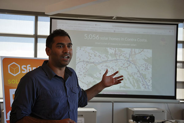 A man speaks and gestures to a slide on a screen behind him.