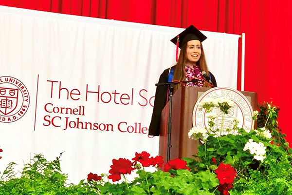 Madison Austrich in a graduation cap and gown standing and speaking at a podium with a banner of The Hotel School and Cornell SC Johnson College of Business in the background.