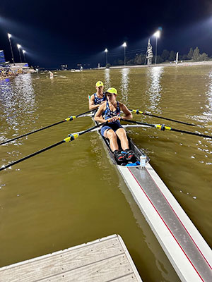 two women, each holding two oars, rowing a long narrow boat.