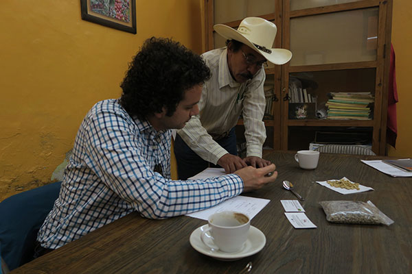 Two men at a table with coffee cups and papers, one standing and one sitting, looking over papers, pointing, and discussing.