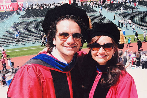 a man and a woman dressed in red and black PhD gowns and hoods, smiling, when bleachers of Cornell's footbal stadium in the background.