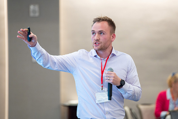 a man standing and speaking into a microphone as he gestures and looks at a screen to his right.