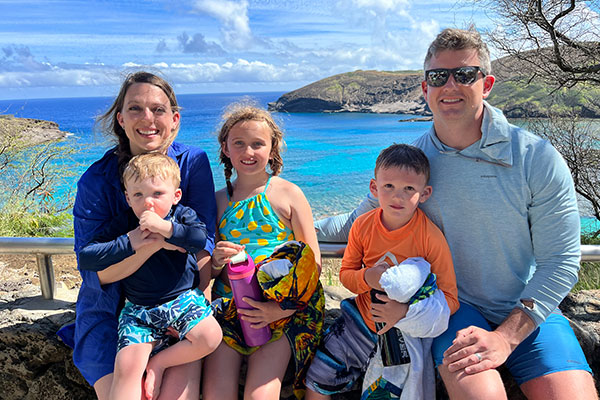 a woman, man, and 3 small children sitting down at a lookout with the ocean and some trees in the background.