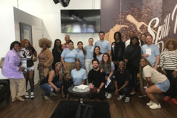 A large group of people posing against a wall at one end of a room and smiling at the camera.