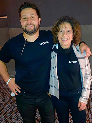 A man and a woman standing side by side, facing the camera, with arms around each other and smiling. Both are wearing black T-shirts with “Tres Leches” on the upper left.