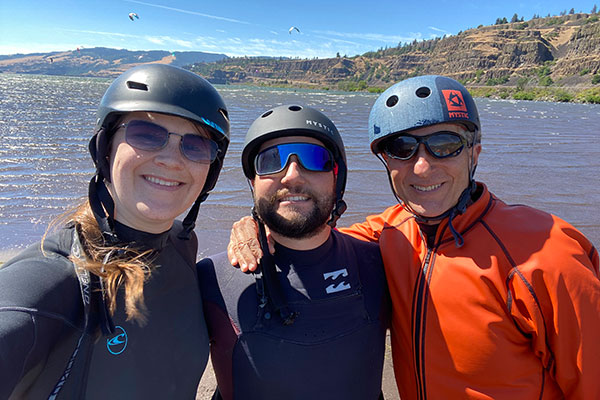 A young man, a young woman, and an older man in the foreground, wearing helmets, with a wide river and cliffs of a river bank and people kitesurfing against a blue sky in the background.