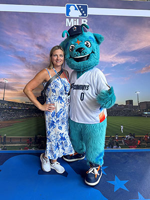 Marianne Boak standing next to a big blue fluffy dog mascot in front of a banner depicting a green baseball field and stands.