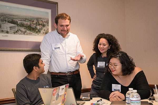 Four students dressed in business casual, two seated and two standing, around a table and talking while looking at a laptop screen.