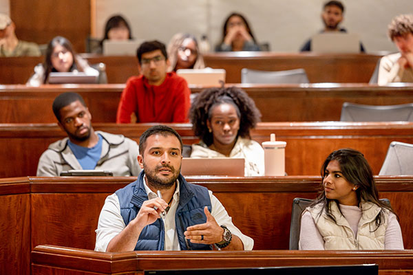 Students in tiered lecture hall seating. One is speaking and gesturing while others look on.