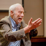 John Marks leaning forward in his chair and gesturing with both hands in the air; his facial expression shows he is passionate about what he is saying.