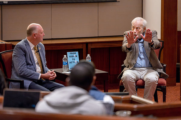 Two men seated in armchairs on a stage, one looking on as the other gestures.