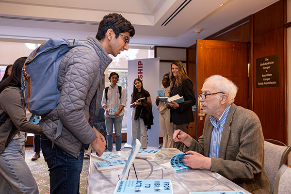 John Marks seated at a table with several copies of his book, about to sign one of the books and looking up at a student who is standing across the table from him and leaning down towards him. Several other students are standing in line in the background, holding copies of his book.