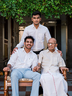 A young man and a much older man sitting on an upholstered wooden bench with a young man standing behind them, his hands on each shoulder. They are outside in a courtyard with foliage from a tree hanging over them.