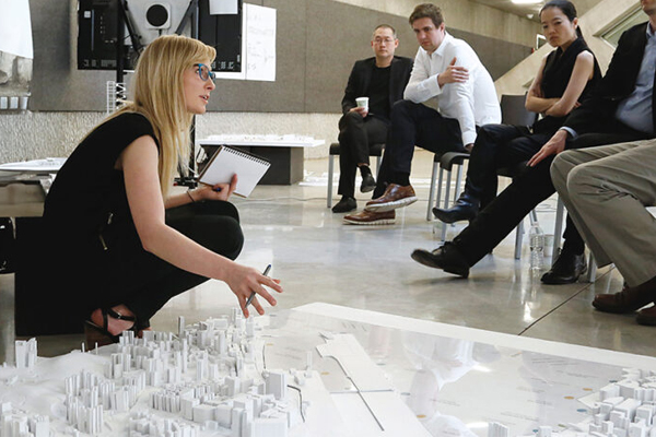 A woman crouches on the floor jestering to a model of a city and speaking in front of people sitting in chairs.