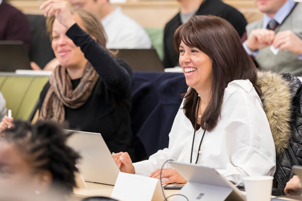 A student smiles and laughs in front of a laptop in a tiered classroom with another student raising her hand in the background.