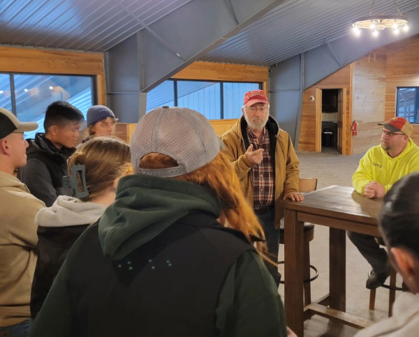 Wayne Knoblauch, wearing a red Cornell baseball cap and tan jacket, speaks to a group of students in a farm building.