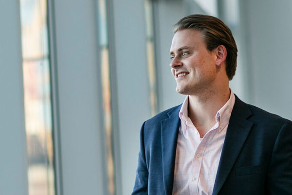 A student in a button up shirt and suit jacket smiles and looks out a wall of windows.