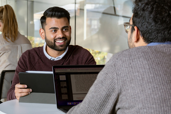 A student in a sweater sits in front of a tablet and smiles at another student in front of a laptop.