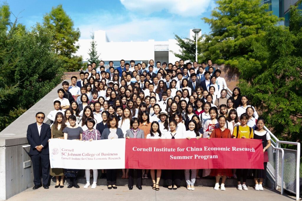 Large group of participants and faculty from the Summer Program standing together outdoors, holding a red banner with program details, in front of a modern building surrounded by greenery.