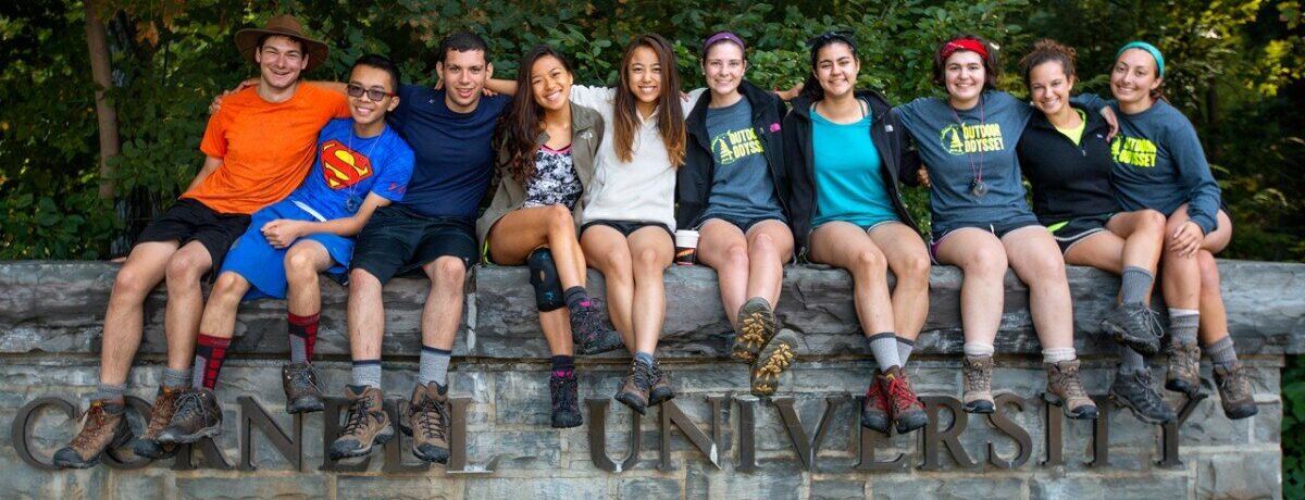 Group of smiling students sitting on a stone wall at Cornell University, surrounded by greenery, representing the collaborative and engaging environment of the Cornell CICER Summer Program for High School Students.