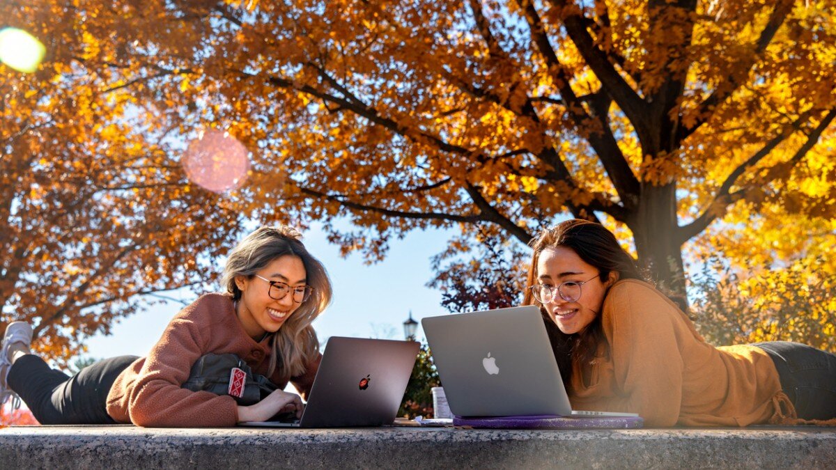 Two students studying with laptops under the vibrant autumn foliage on the Cornell University campus, enjoying a sunny day.