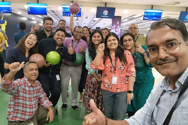 A group of men and women posing for a photo in a bowling alley, smiling, with several holding up bowling balls.