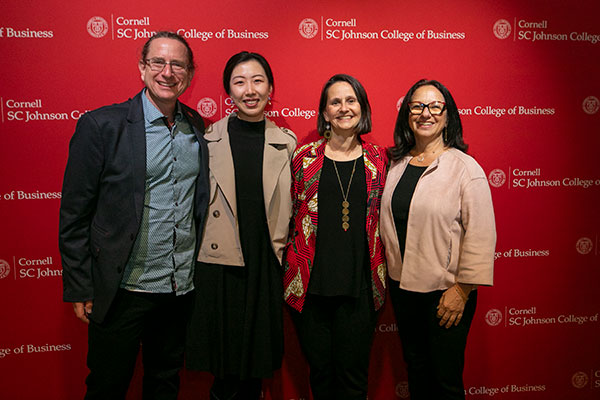  One man and 3 women standing side by side in front of a red backdrop with the Cornell logo and text that reads: Cornell SC Johnson College of Business.