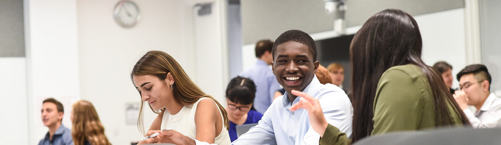 Students sit at a desktop in a tiered classroom talking and smiling.