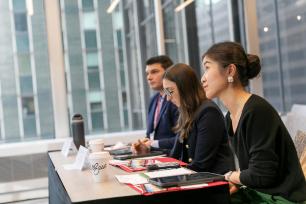 Three people sit at a long table with notebooks and computers, looking intently at a presentation.