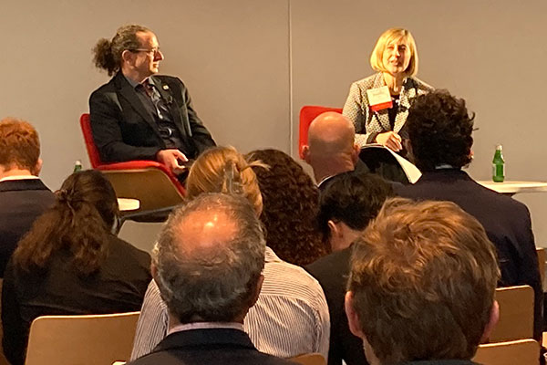 a man and a woman seatd on stage; the woman is speaking into a microphone. The backs of the audience are in the foreground.