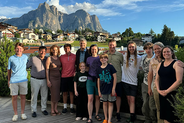 More than a dozen adults and children dressed in summer clothes posing for a photo with an alpine village, rock-faced mountains, and bright blue sky in the background.