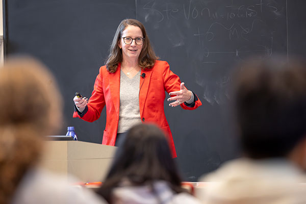 A woman standing at a podium in front of a classroom of students, speaking, smiling and gesturing with both arms.