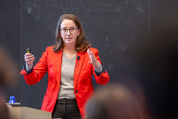 A woman standing at a podium in front of a classroom of students, speaking, smiling and gesturing with both arms.