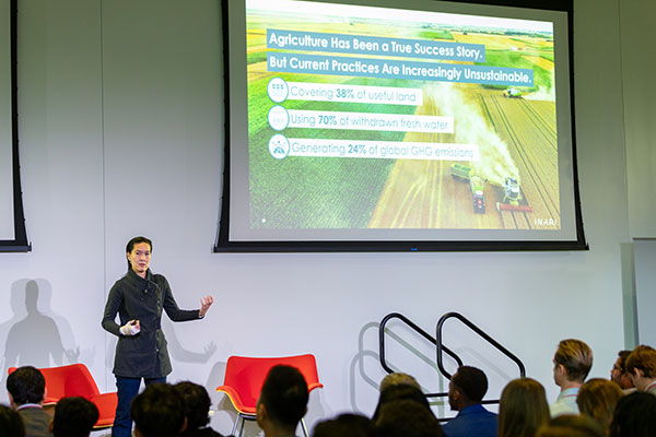 A woman on a stage speaking and gesturing. Behind her is a screen showing the statistics cited in the text: The agricultural industry takes up 38 percent of the world’s arable land, uses 70 percent of its freshwater, and is responsible for nearly 25 percent of greenhouse gas emissions.