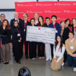 Winners of the competition holding a large check, surrounded by other participants. Organizers and funders are standing behind them; a red Cornell banner appears in the background.