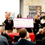 Students stand with a bear mascot and an oversized check in front of an audience as gold confetti falls.