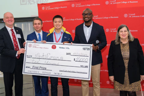 Roy Wang holds the first-prize check with Carlos Bernos, Osagie Oigiagbe, Dean Andrew Karolyi, and Lourdes Casanova on the conference stage with a red Cornell banner in the background.