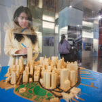 Female student takes notes while observing a wooden architectural model of a city displayed in a glass case. The background features an exhibition space with displays, informational panels.