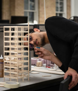 A male student leans in closely while working on a detailed wooden architectural model of a multi-story building. He is carefully adjusting a small piece with his hands. The workspace is filled with design materials, including glue, wooden pieces, and a desk lamp, in a studio setting.