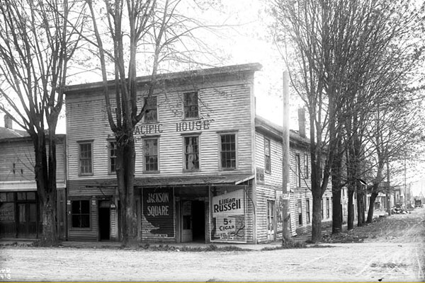 black and white photo of a 3-story wood, clapboard building on a street corner.