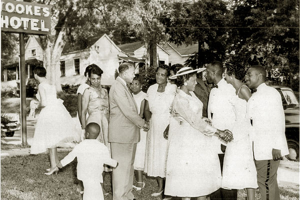 Wedding buests standing on the lawn in a receiving line being greeted by and shaking hands with by a man and a woman. In the background are other houses and a tall neon sign, "Tookes Hotel."