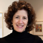 Headshot of a woman with brown curly hair standing in an art gallery.