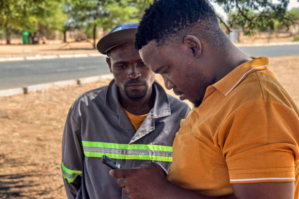 Two men look at a mobile phone near a road in a dry landscape.