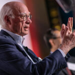 A close-up photo of a man’s head and hands as he gestures with his hands and speaks.