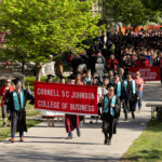 Commencement processional of students in the SC Johnson College of Business dressed in graduation regalia and holding a college banner.