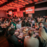 a large banquet hall filled with people seated at round tables and with two large red banners with the text Big Red Bash hanging on either side of the stage.